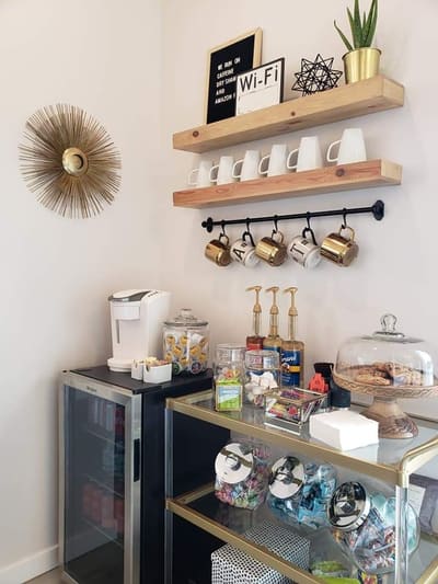 The complimentary refreshment bar at AT.hair & co.: a brass and glass cart with a coffee maker, flavored syrup pumps, cookies under a glass cloche, candy jars, a beverage fridge, and mugs on floating wood shelves.