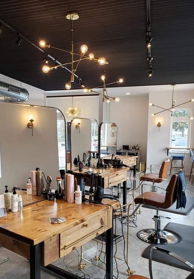The styling floor at AT.hair & co.: reclaimed-wood stations with professional product and hot tools, arched brass mirrors, brass sconces and sputnik chandeliers, a black shiplap ceiling, and cognac leather styling chairs.