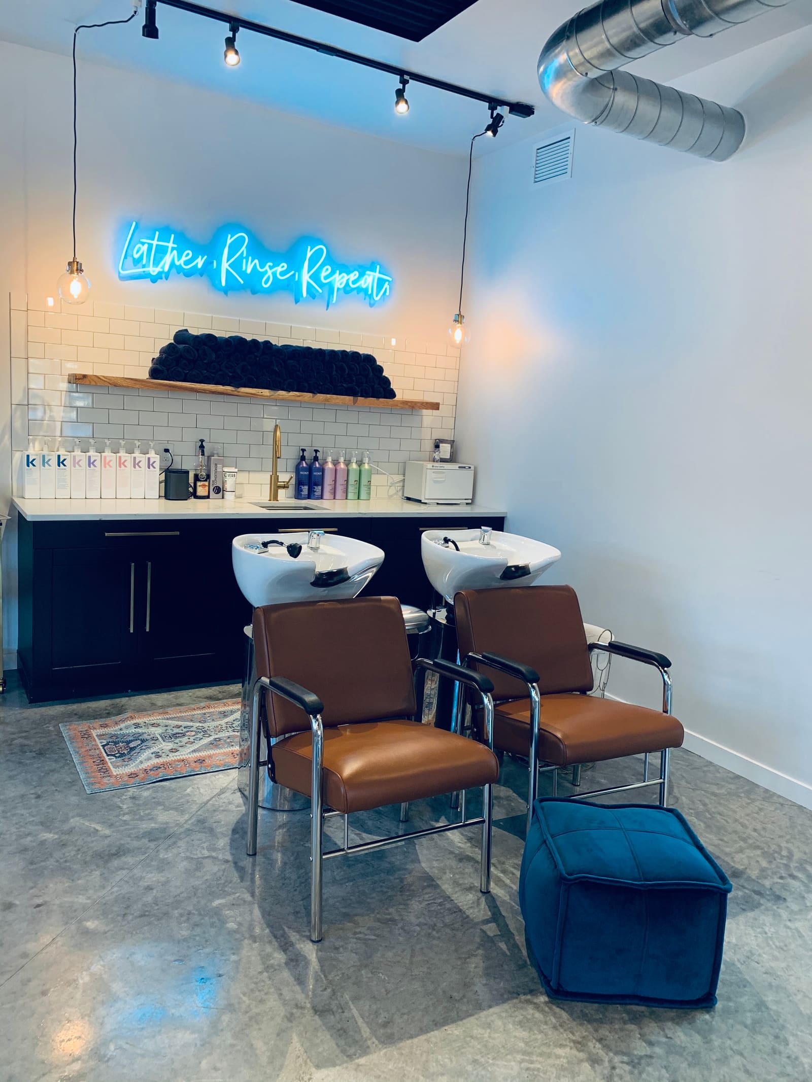 The shampoo area at AT.hair & co.: two white bowls with cognac leather reclining chairs, white subway tile, rolled black towels on a floating wood shelf, a towel warmer, and a blue neon script sign reading Lather, Rinse, Repeat.